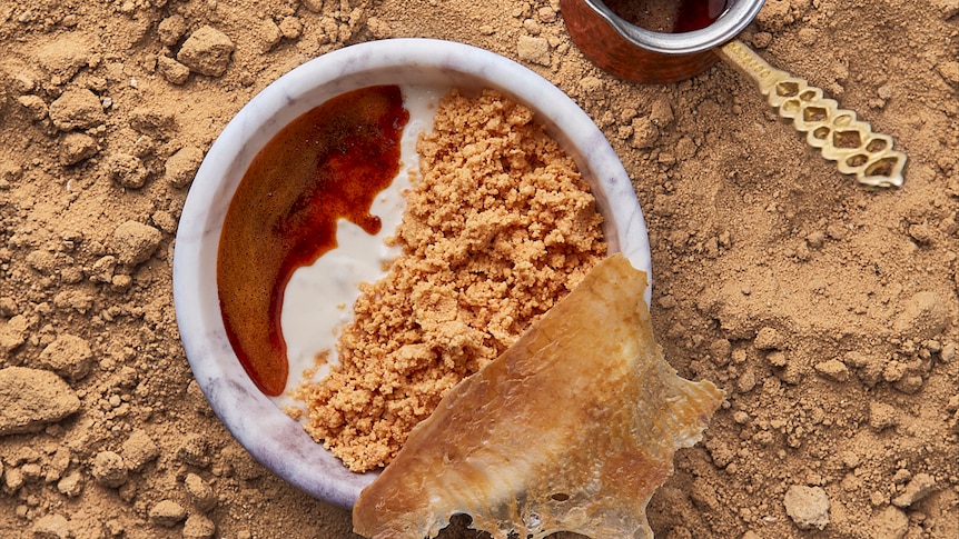 A poached eggs and yoghurt dish in a ramekin with a small decorative pot of caramel, set on sand.