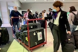 A woman and man wearing face masks wheel a trolley with bags and boxes through an airport.