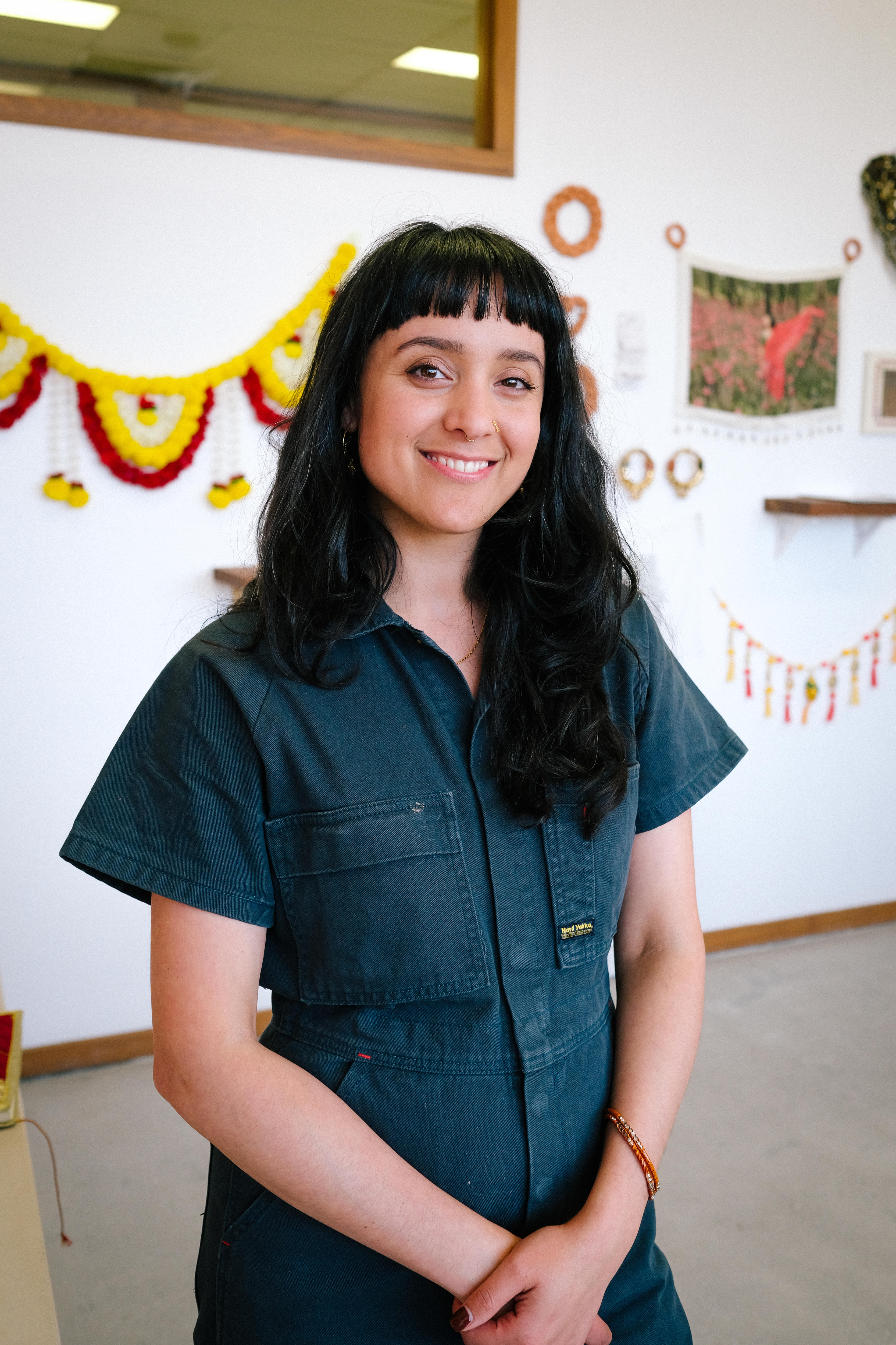 Artist Monica Rani Rudhar smiles warmly at the camera in her studio — she has thick black hair with a fringe.