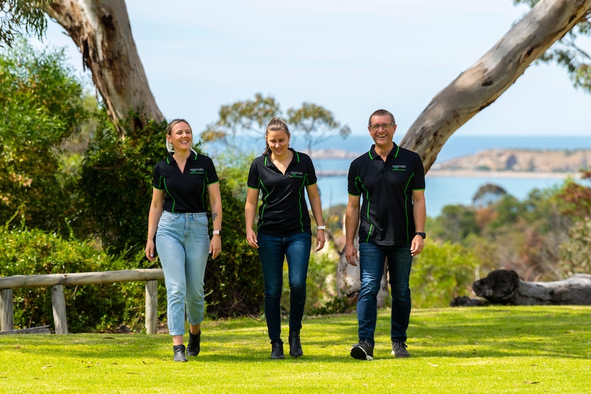 Two women and a man all wearing black uniform polo shirts walk across the grass with trees and the ocean visible behind.