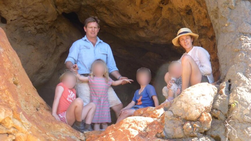 A man and woman pose in a rocky cave opening with four children. The children's faces are obscured.