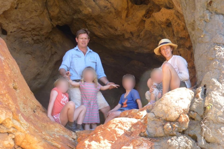 A man and woman pose in a rocky cave opening with four children. The children's faces are obscured.