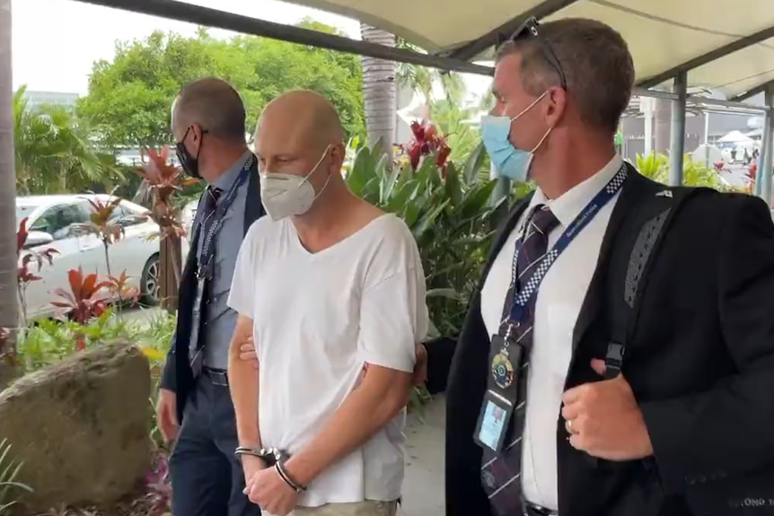 a bald man in handcuffs flanked by two police detectives wearing suits and ties