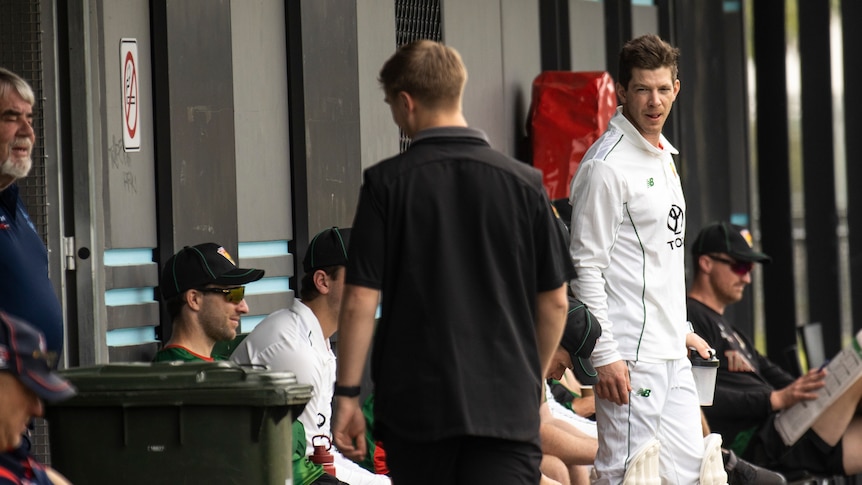 Man in cricket whites and pads stands in front of team mates waiting to bat. 