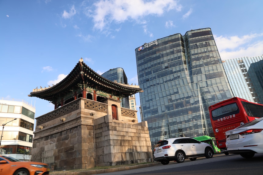 An old Korean structure in foregound with modern, glass high rise in background. Both alongside a busy road filled with vehicles