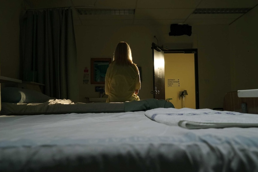 Woman with blonde hair sits on hospital bed in the dark facing an open door