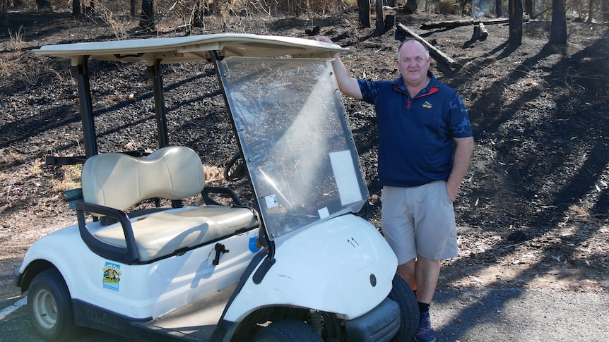 A man stands next to a golf buggy in front of burnt out trees 