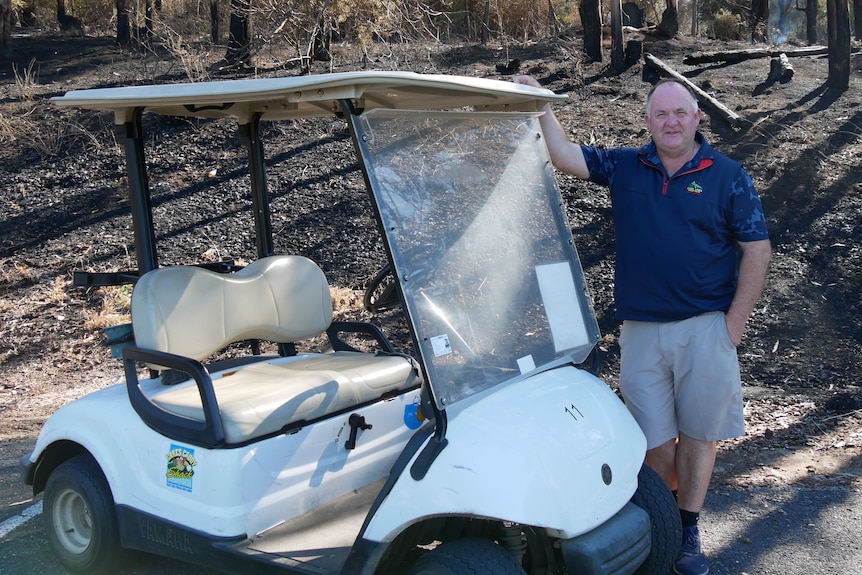 A man stands next to a golf buggy in front of burnt out trees 
