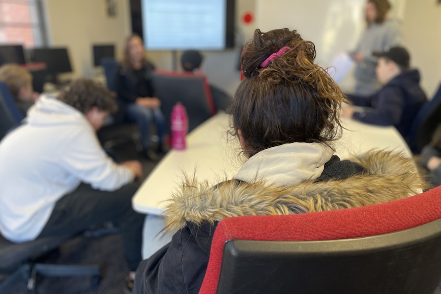 Teenagers, whose faces we cannot see, sit at desks in a classroom.