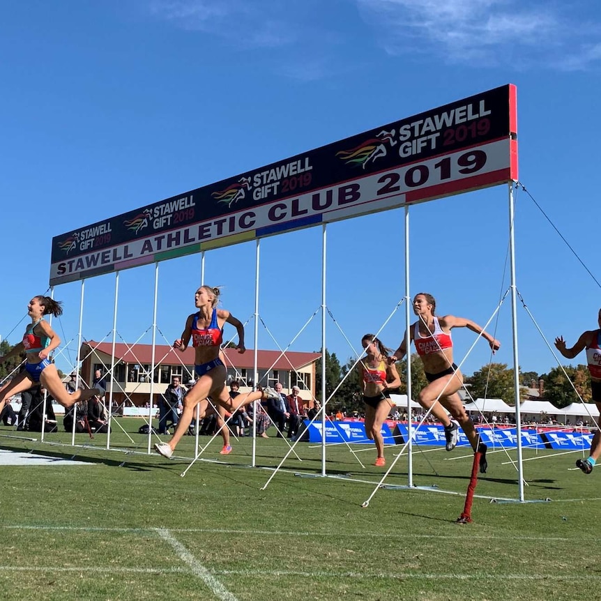 A group of women sprint over the finish line at the Stawell Gift in front of a blue sky.