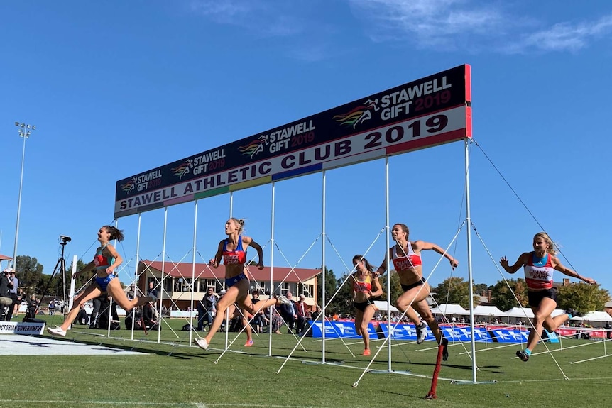 A group of women sprint over the finish line at the Stawell Gift in front of a blue sky.