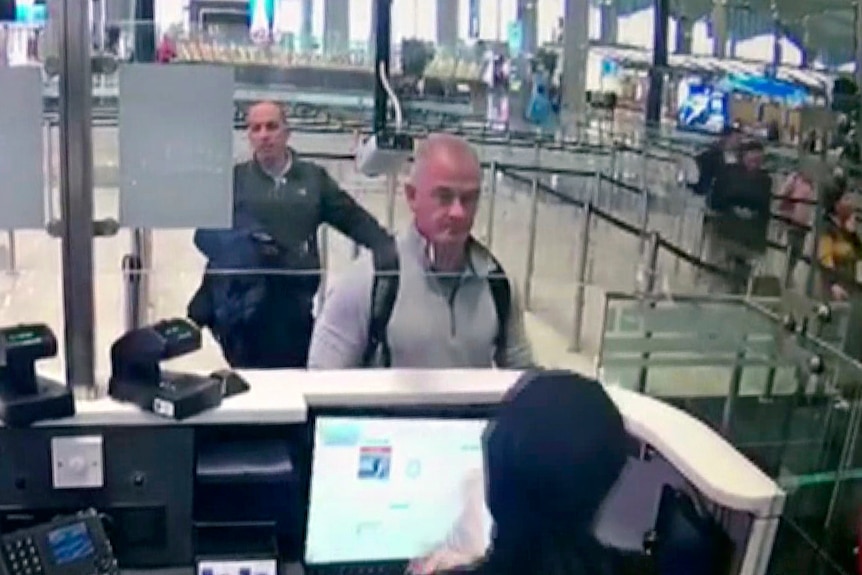 An image from a security camera shows two men in casual clothes approching a passport control desk at an airport.