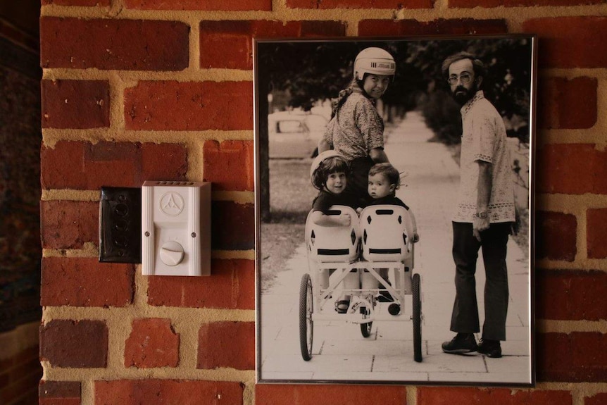 A woman wearing a bike helmet looks at the camera with two children sitting on a bike behind her, and her husband nearby.