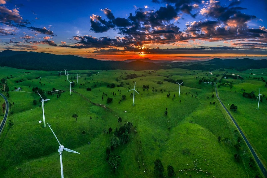 Wide angle aerial view of wind turbines in the foreground with the sun rising in the far distance.