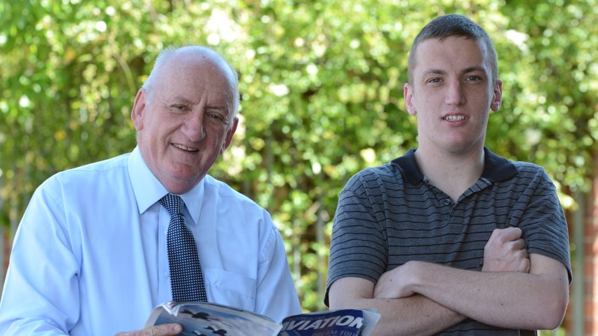 Elderly man in shirt and tie smiles holding an aviation magazine next to man in stripy shirt with crossed arms.