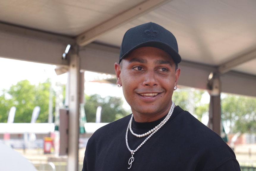 a young aboriginal man smiling, wearing a black cap, t-shirt and silver necklaces