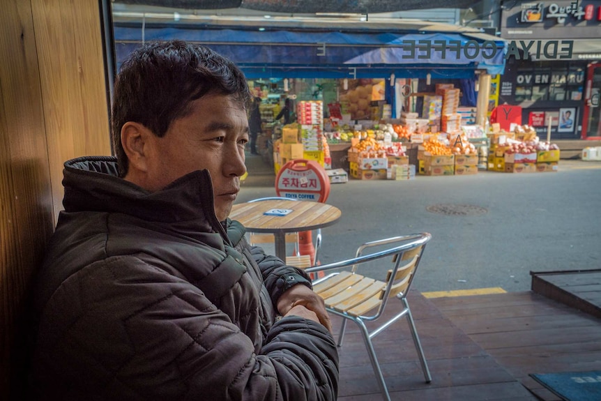 A middle aged Asian man with a forlorn expression looks outside from an Ediya Coffee shop.