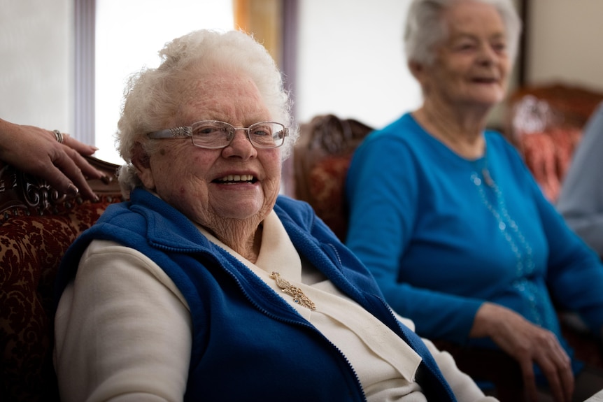 An older woman with glasses smiling while sitting at a table with other older women.