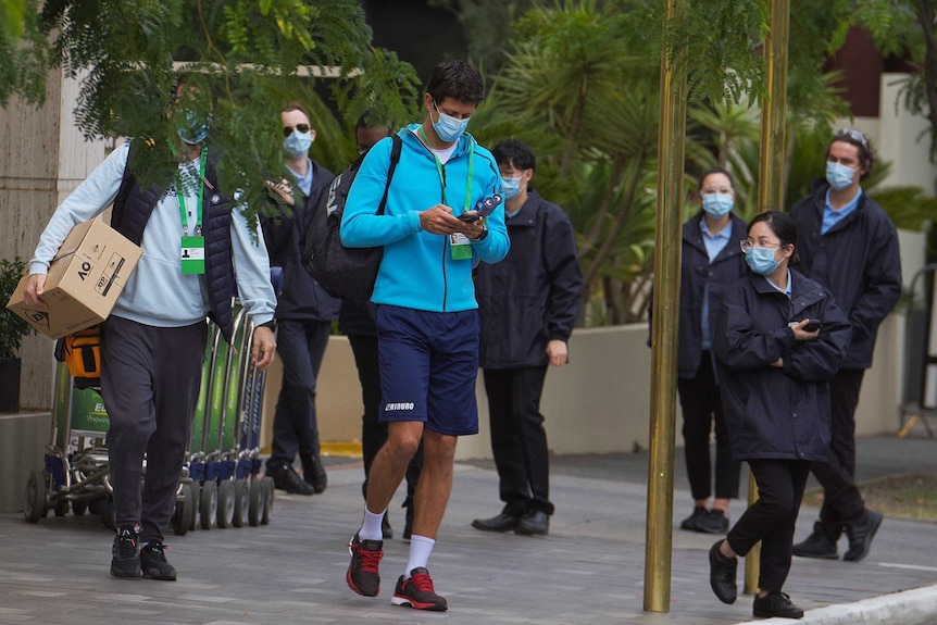 A tennis player wearing a mask is escorted by hotel quarantine staff.