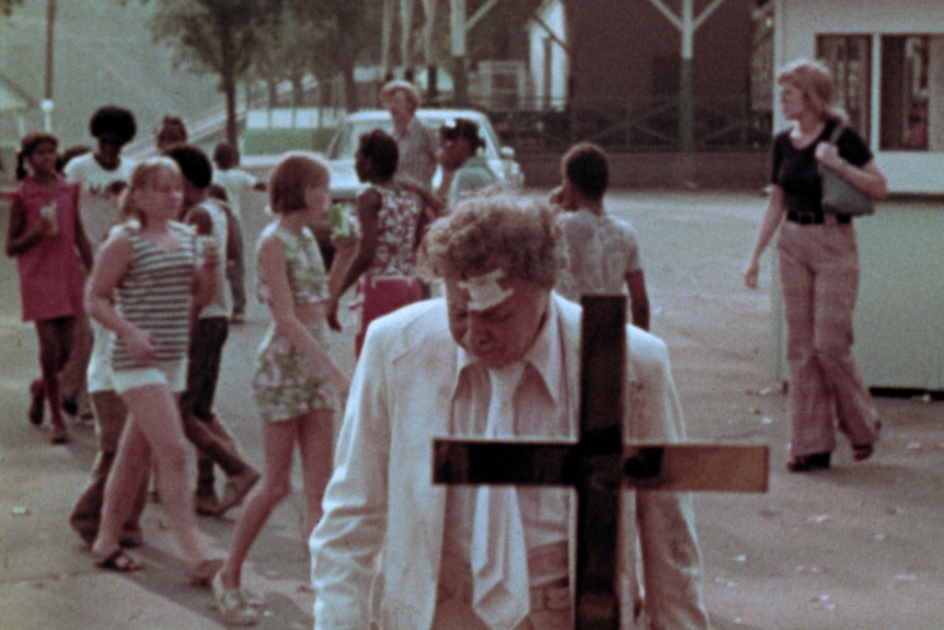 Film still of a battered Lincoln Mazel standing in front of a cross, while people mill around behind him in The Amusement Park