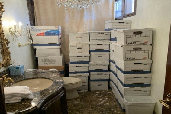 A stack of cardboard boxes sit on the floor of a bathroom between a toilet and a shower.