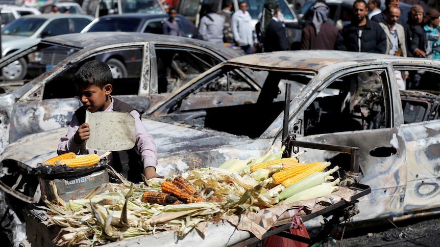 A boy sells grilled corn near damaged cars.