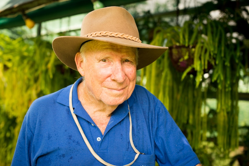 A man wearing a hat smiles in front of a series of hanging ferns.