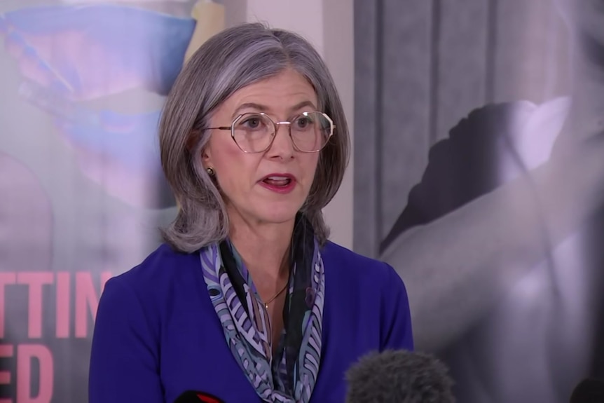 A women in a purple blazer and glasses speaking to microphones