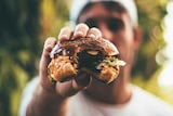 A man holds a hamburger towards the camera, offering a bite.