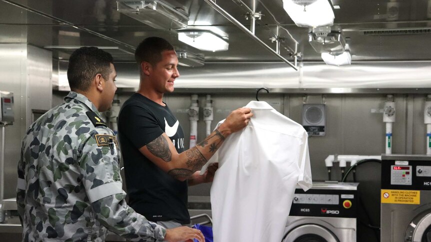 Two men stand in HMAS Brisbane's laundry, one holding a shirt.