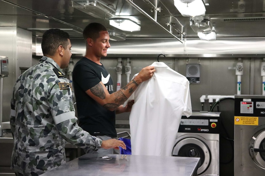 Two men stand in HMAS Brisbane's laundry, one holding a shirt.