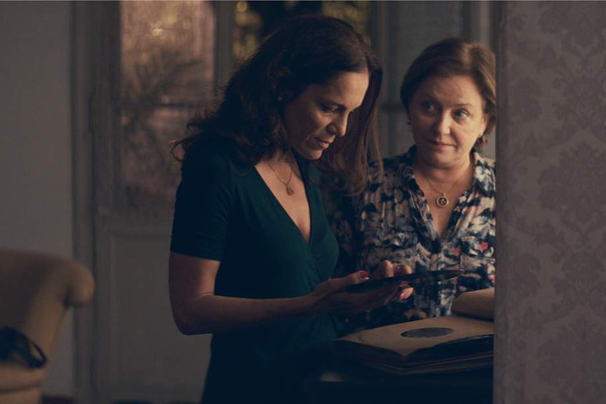 A 40-ish woman and a 60-ish woman in a living room, looking at a record as they stand close together.