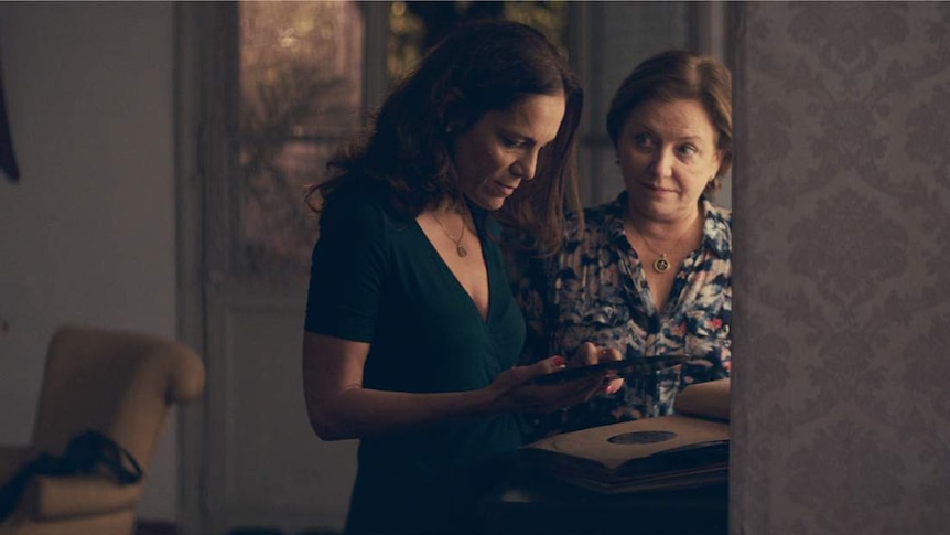 A 40-ish woman and a 60-ish woman in a living room, looking at a record as they stand close together.