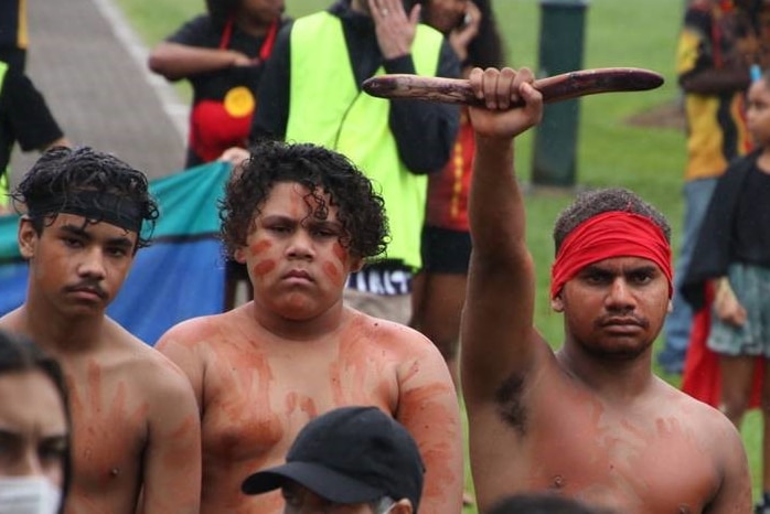 Four young men with ochre on their torsos stand in a crowd of people. One of the men wears a red headband and raises a wood item