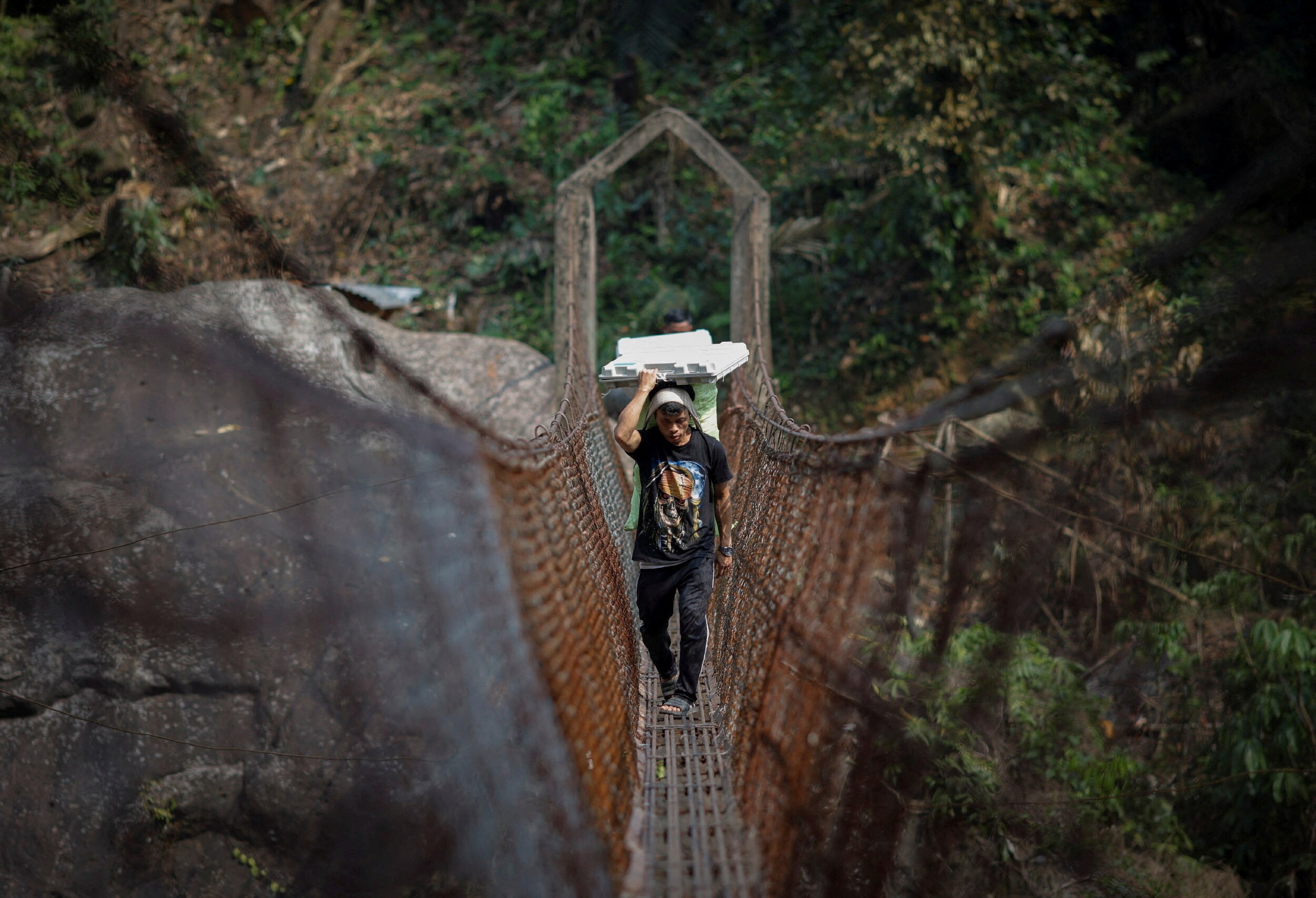 A man carries a white package on his head as he crosses a suspension bridge 