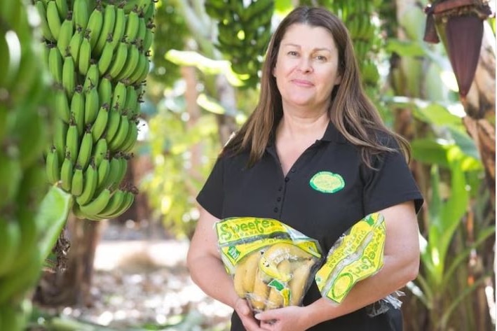 A woman standing in front of banana trees holding packets of bananas