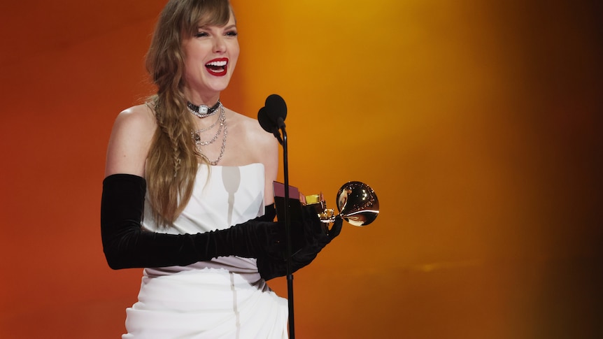 Taylor Swift holding a Grammy and smiling widely wearing a white sleeveless evening gown and long black gloves