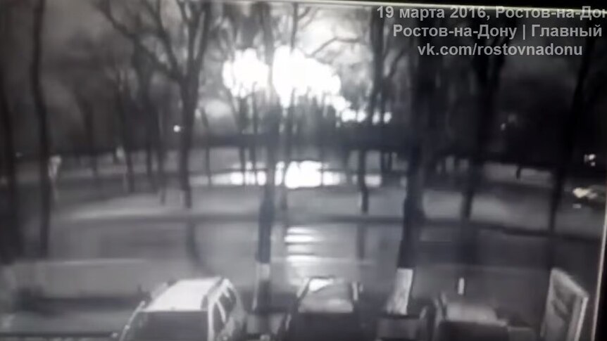An explosion is visible in the background of trees and a line of cars, in black and white CCTV footage