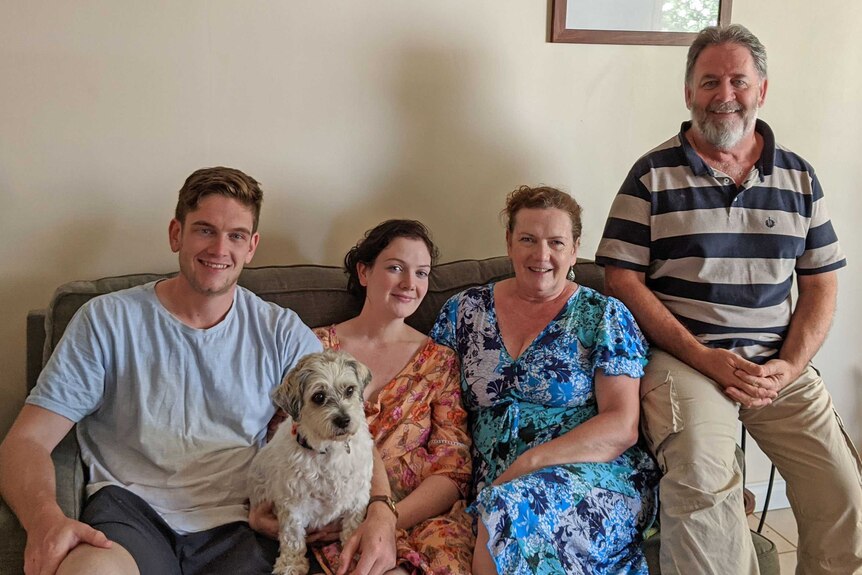 A family of two adult children and two parents sitting on a couch in a family home with a pet dog on their lap.