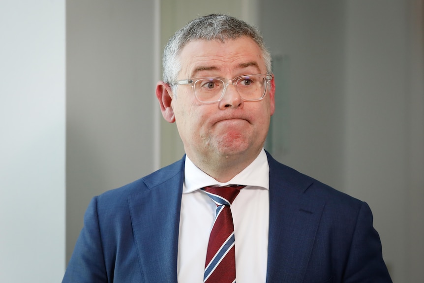 A man wearing a tie and suit. He has grey hair and clear-rimmed glasses and a downward smile with his lips pulled inwards.