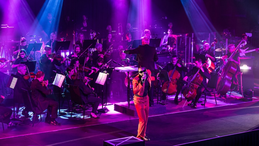 Genesis Owusu wears a red suit and sings into a microphone. The Brisbane Symphony Orchestra plays behind him.