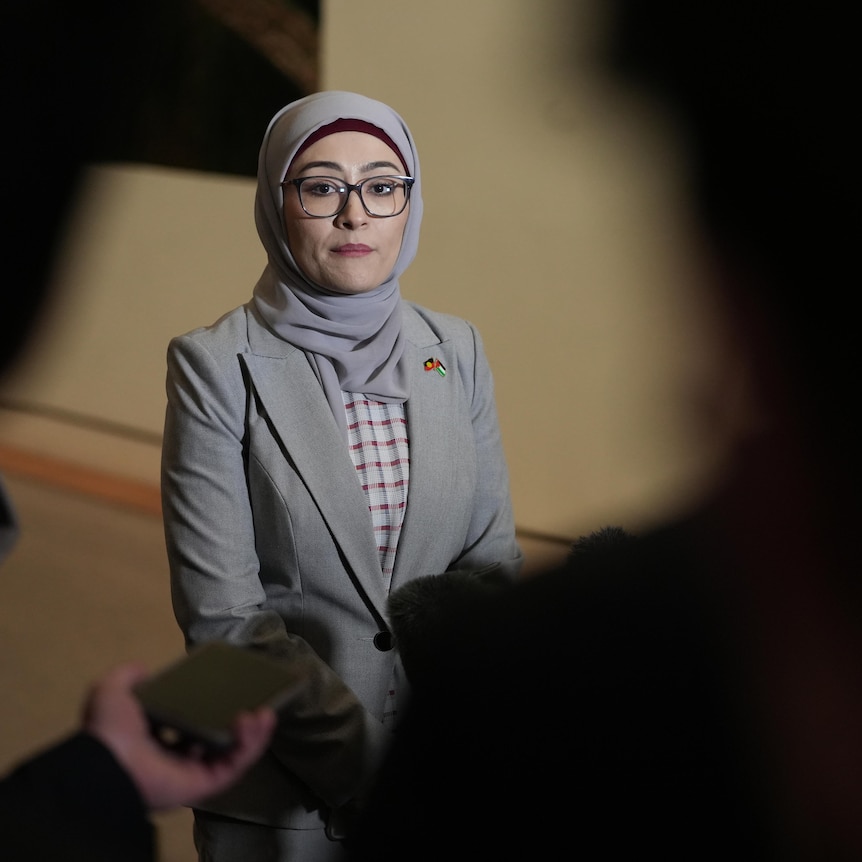 A woman wearing a pale grey jacket and hijab, and glasses.