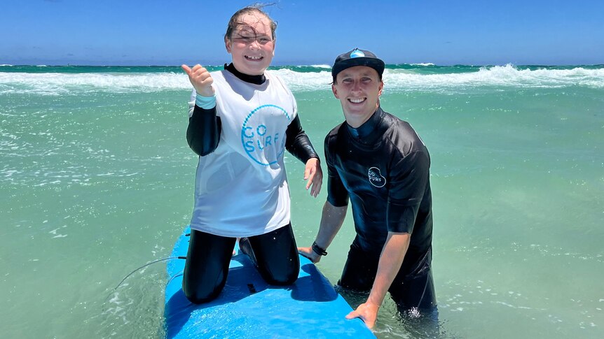 Young girl on a surfboard in the water next to a surf instructor in a wetsuit.