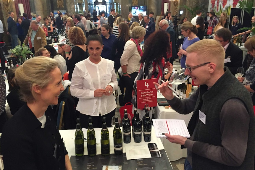 A wine-maker, taster and attendant stand in the foreground as crowds stand behind them.