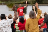 A man wearing a red shirt with his hand in the air in front of a group of teachers. 