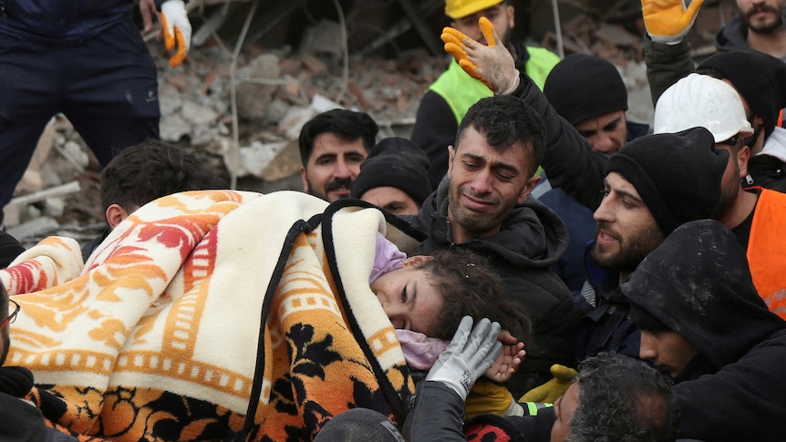 People carry a girl who is laying down covered in a patterned blanket. 