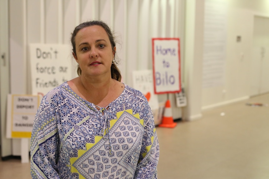 A serious middle-aged woman, hair tied back, wearing white and blue blouse stands in from of signs saying Home to Bilo.