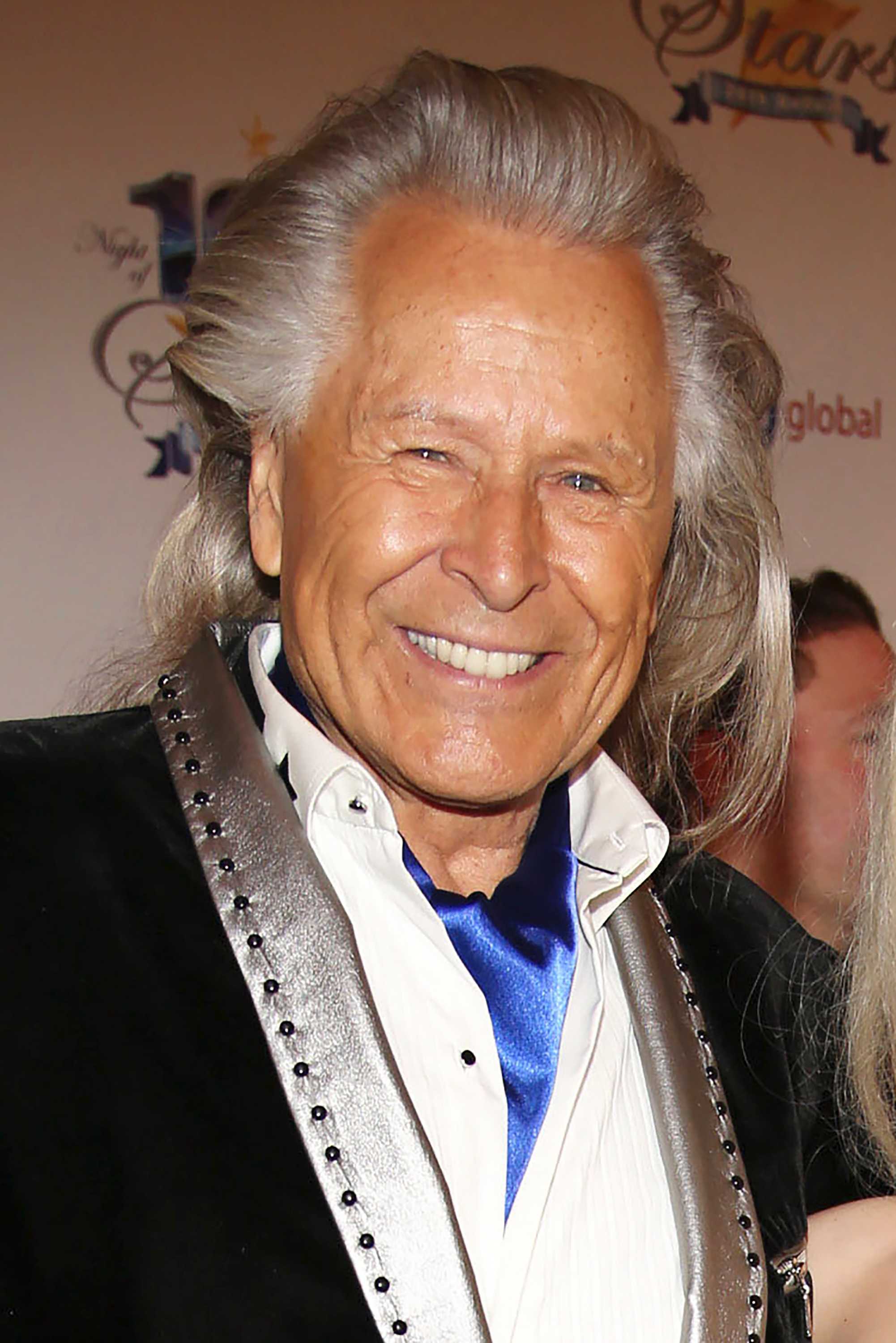 Elderly man with long, grey hair wears black suit with silver collar and white shirt