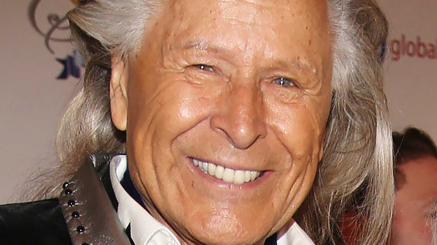 Elderly man with long, grey hair wears black suit with silver collar and white shirt