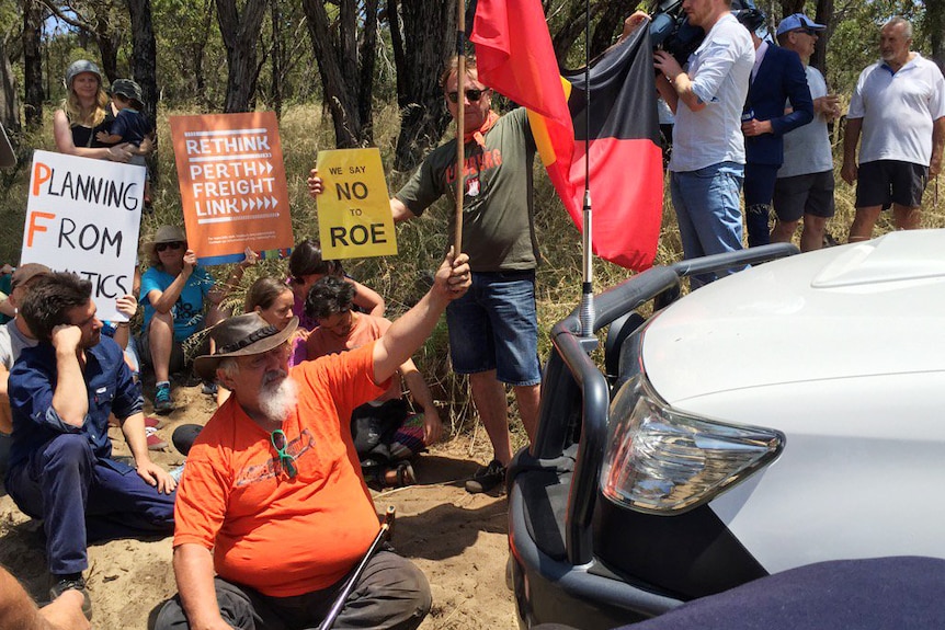 Protesters carring placards and an Aboriginal flag sit in front of a Main Roads ute.